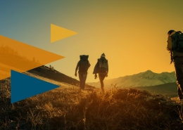 Three hikers with backpacks walk across a sunlit hillside at sunset toward distant mountains with a warm gradient sky.