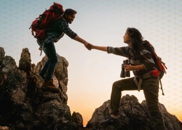 Two hikers on a rocky ridge reach out to shake hands at sunrise, with backpacks and gear.